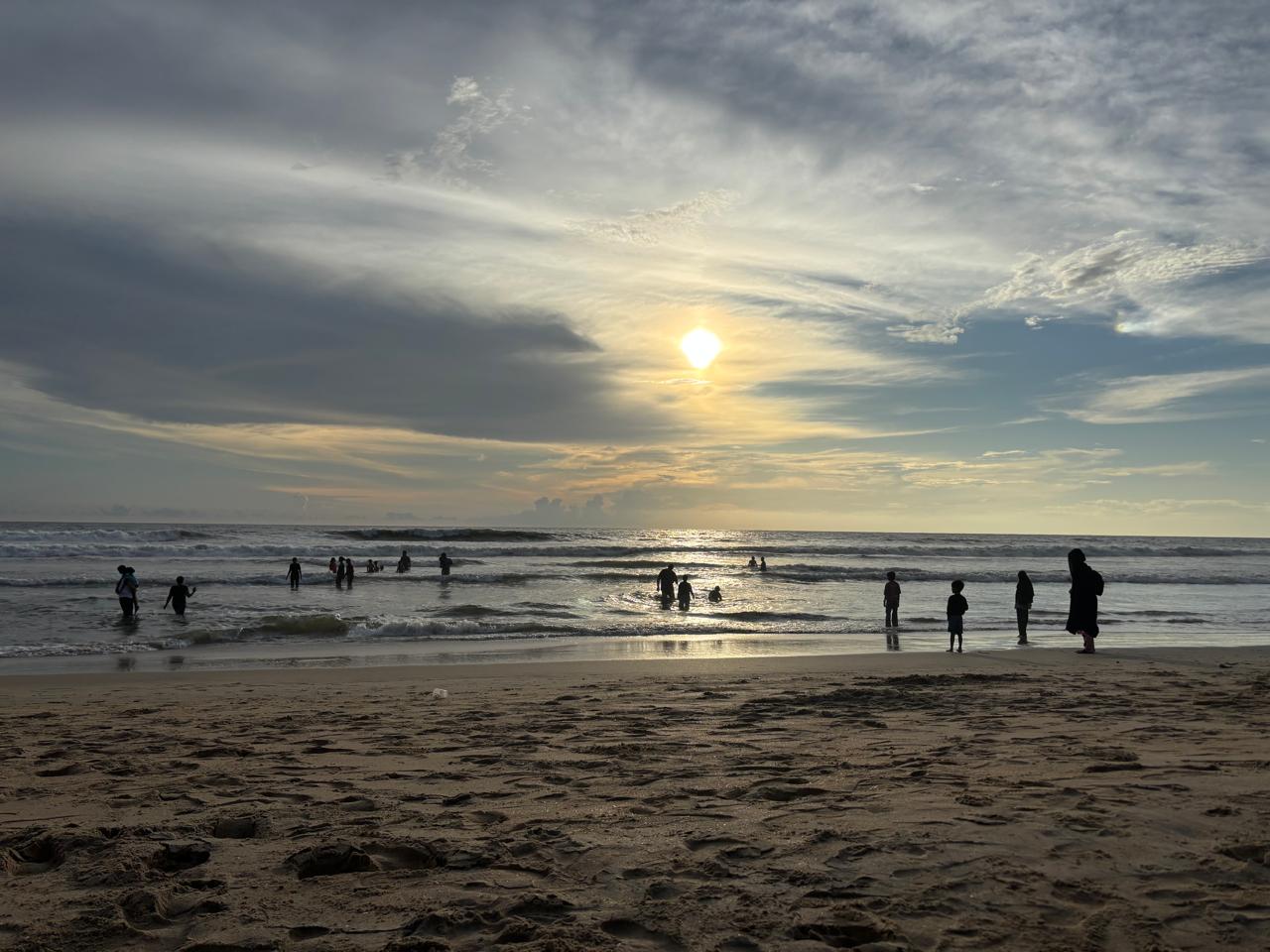 Evening sky over the Arabian Sea, sun breaking through cloud, figures wading at the shoreline