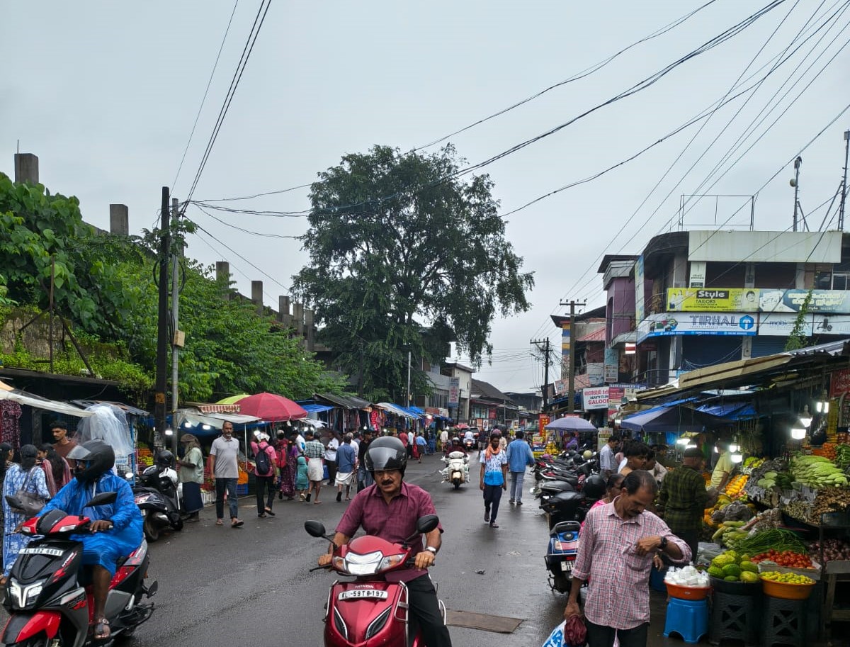 A busy street market in Kannur town on a grey monsoon morning, shoppers and mopeds moving past stalls of fruit, vegetables and clothing