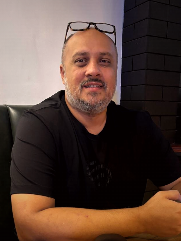 Portrait of Saqib Chinoy, glasses pushed up, smiling in a café booth