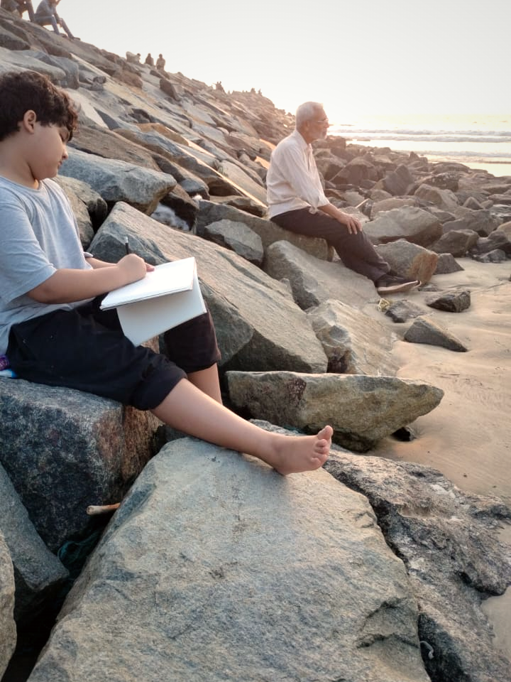 A child sketching in a notebook on the rocks while an elder sits looking out to the sea at golden hour
