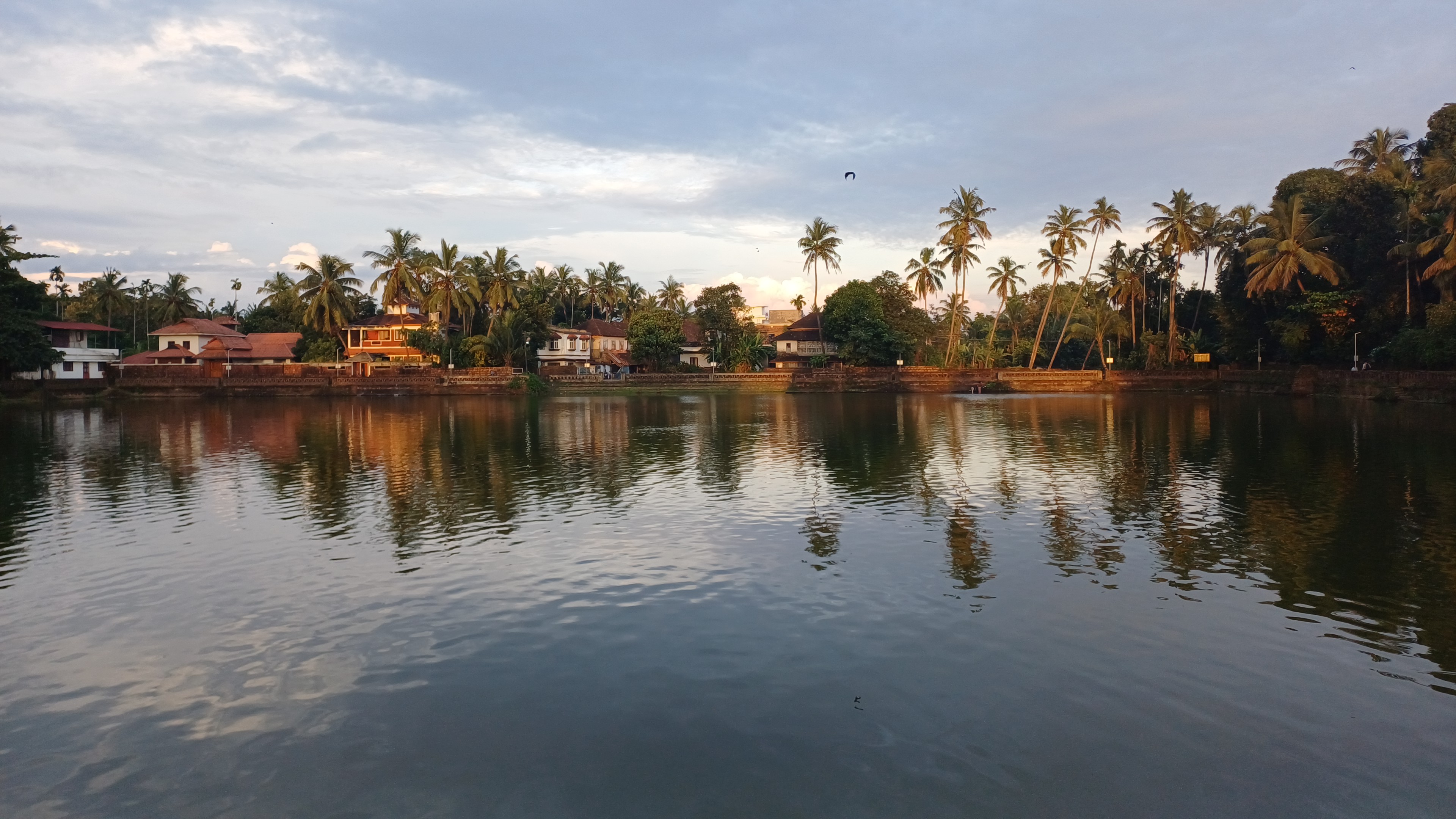 A temple pond at dusk, old tiled houses and palms reflected in the water under a soft sky